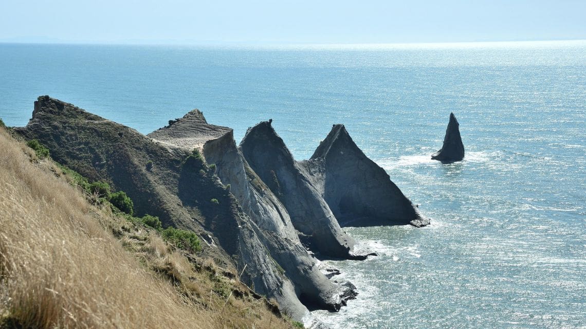 Coastal landscape with rugged, jagged cliffs extending into a calm blue ocean. Grassy slopes top the cliffs, evoking a serene and majestic scene.