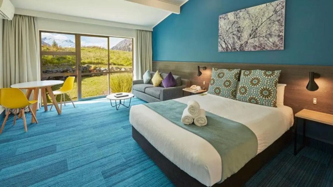 Modern hotel room with blue tones features a queen-sized bed, sofa with colorful pillows, and a table with yellow chairs. Large window shows a grassy landscape.