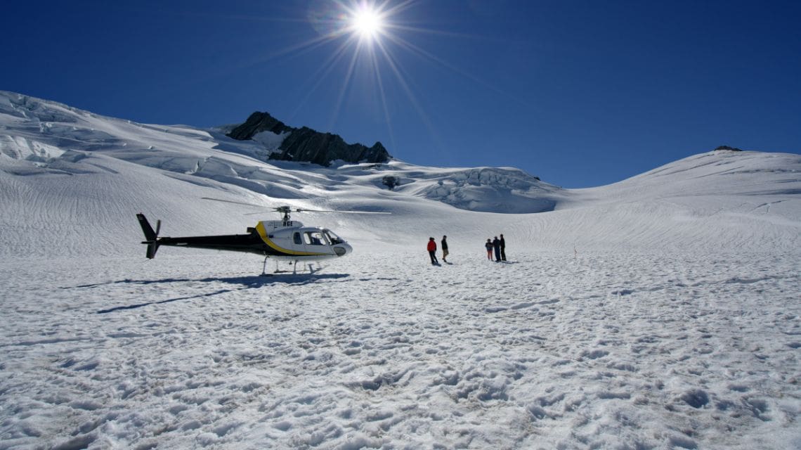 A helicopter sits on a vast snow-covered glacier under a clear blue sky and bright sun. Nearby, five people stand in conversation, conveying exploration and adventure.