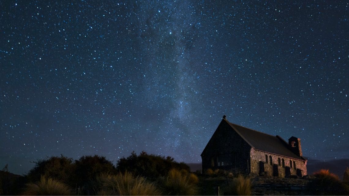 A stone church stands under a vast, star-filled night sky, with the Milky Way prominently visible. The scene evokes tranquility and wonder.