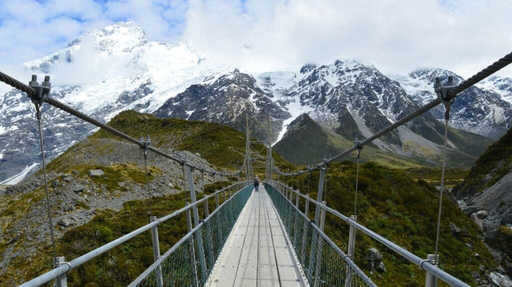 A single person walks across a suspension bridge surrounded by lush greenery, with snow-capped mountains in the background under a partly cloudy sky.