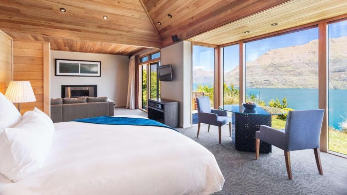 Luxurious bedroom with a wooden ceiling, large windows showcasing a serene lake view. Includes a comfy bed, seating area, and a cozy, relaxed atmosphere.