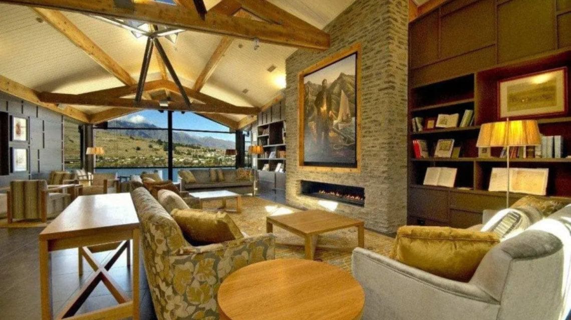 Spacious, elegant lobby with exposed beams and large windows showcasing a mountain view. Features cozy sofas, modern fireplace, and shelves of books.