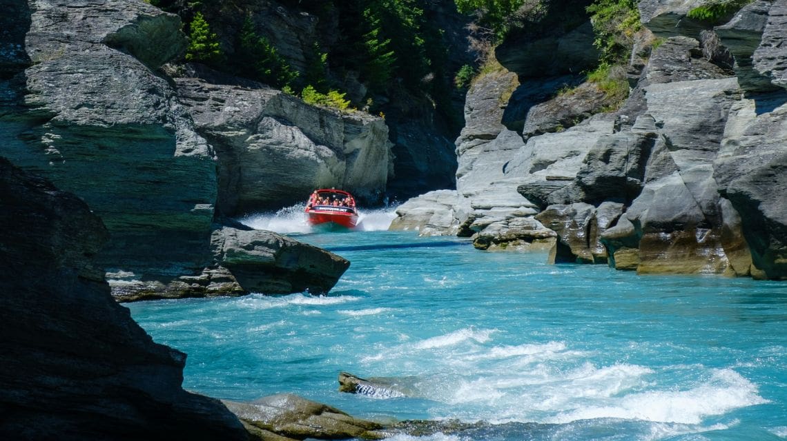 A red speedboat navigates through a narrow canyon with vibrant turquoise water, surrounded by rugged gray rocks and lush green trees under bright sunlight.