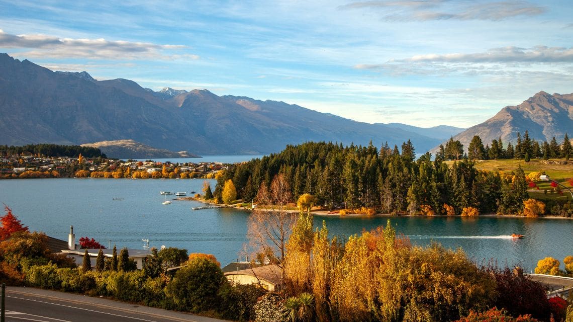Scenic lake view with autumnal trees, vibrant foliage, and distant mountains under a blue sky. A peaceful, picturesque landscape with calm water.