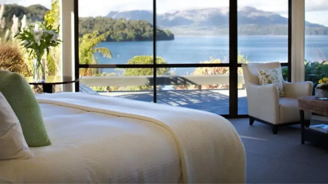 A luxurious bedroom with a plush bed and armchair overlooks a serene lake and mountains through large windows. The scene conveys tranquility and natural beauty.