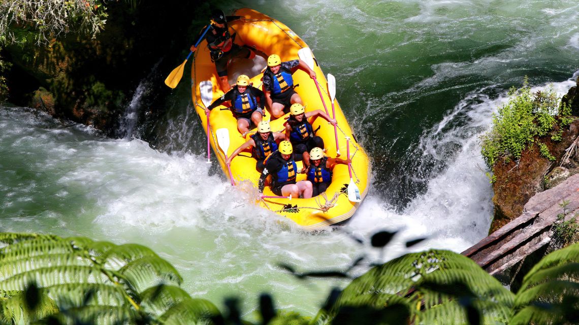 A group of smiling rafters wearing helmets and life jackets navigate through white-water rapids in a bright yellow raft, surrounded by lush greenery.
