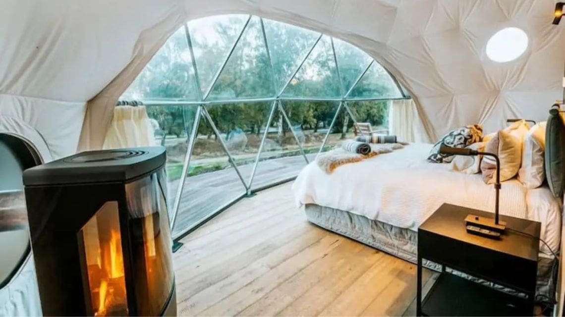 A cozy room inside a geodesic dome with a large window. Features a comfortable bed, wooden floor, modern fireplace, and a scenic woodland view.