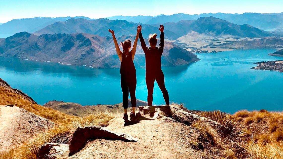Two women on top of a mountain facing away from the camera towards the lake below and mountain range in the background