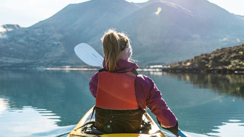 A woman in a yellow kayak, paddling on a lake with a rugged mountain range in the background