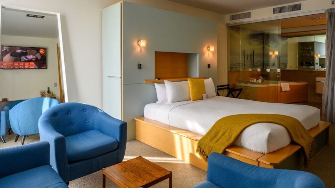 Image of hotel room, large bed with orange blanket. Blue chair next to the bed.