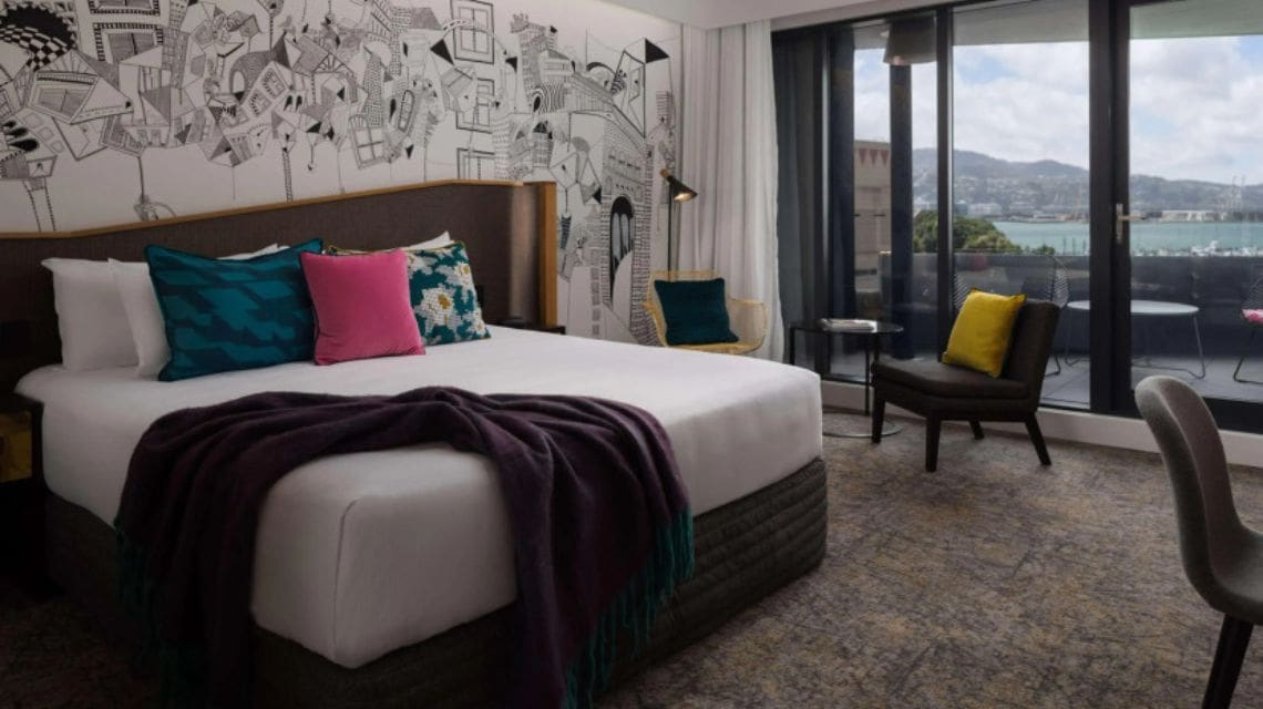 Stylish hotel room with a modern mural behind the bed, colorful cushions, a dark throw blanket, and a balcony view of a bay and hills. Tranquil and inviting.