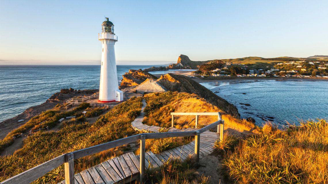 Lighthouse on a rocky cliff at sunrise, overlooking a calm ocean and a small coastal town. Wooden paths and golden grasses create a serene scene.