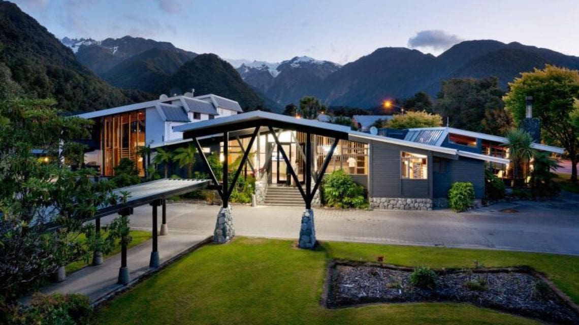 Modern lodge with sloped roofs set against a backdrop of lush mountains and snow-capped peaks at dusk. Warm lighting emits a welcoming ambiance.