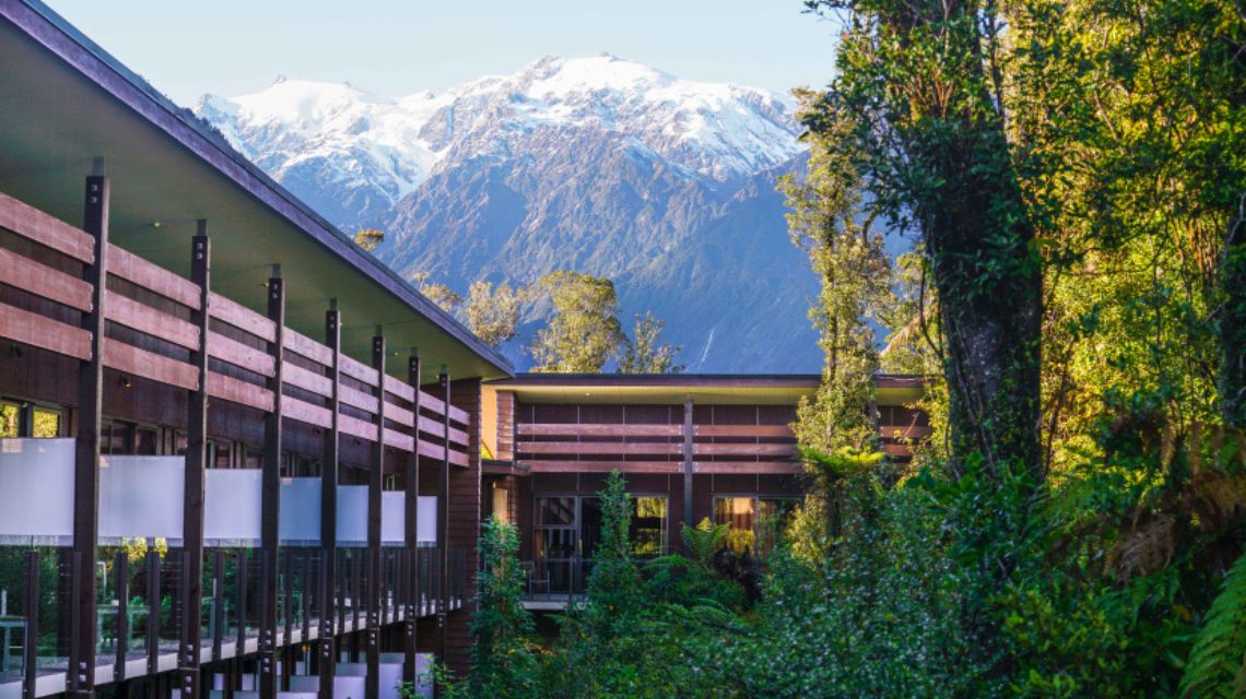 Modern building surrounded by lush greenery, with a backdrop of snow-capped mountains under a clear blue sky, conveying a serene and majestic atmosphere.