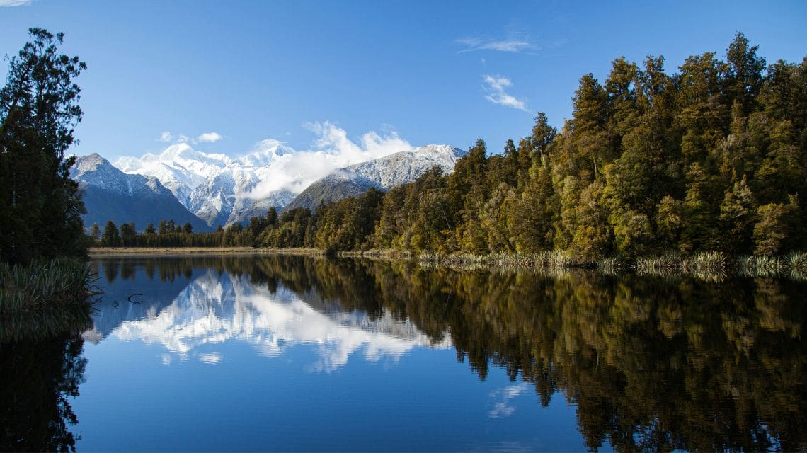 A serene lake reflects snowy mountains and a lush, green forest under a clear blue sky, conveying tranquility and natural beauty.