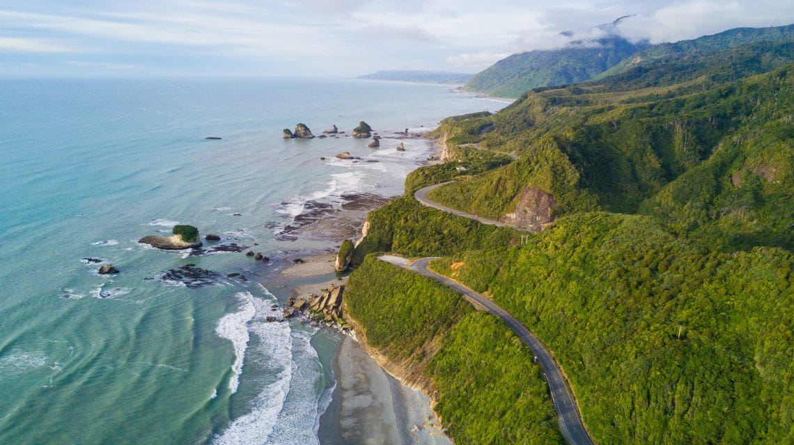 A scenic coastal road winds through lush green hills beside a vast, turquoise ocean. Rocky formations dot the shoreline under a partly cloudy sky.