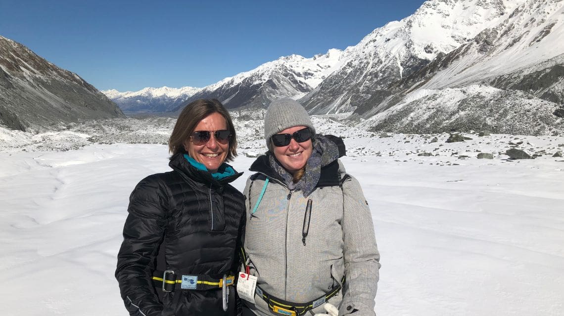 Two women in winter clothes on a glacier with rugged snow capped mountains in the background