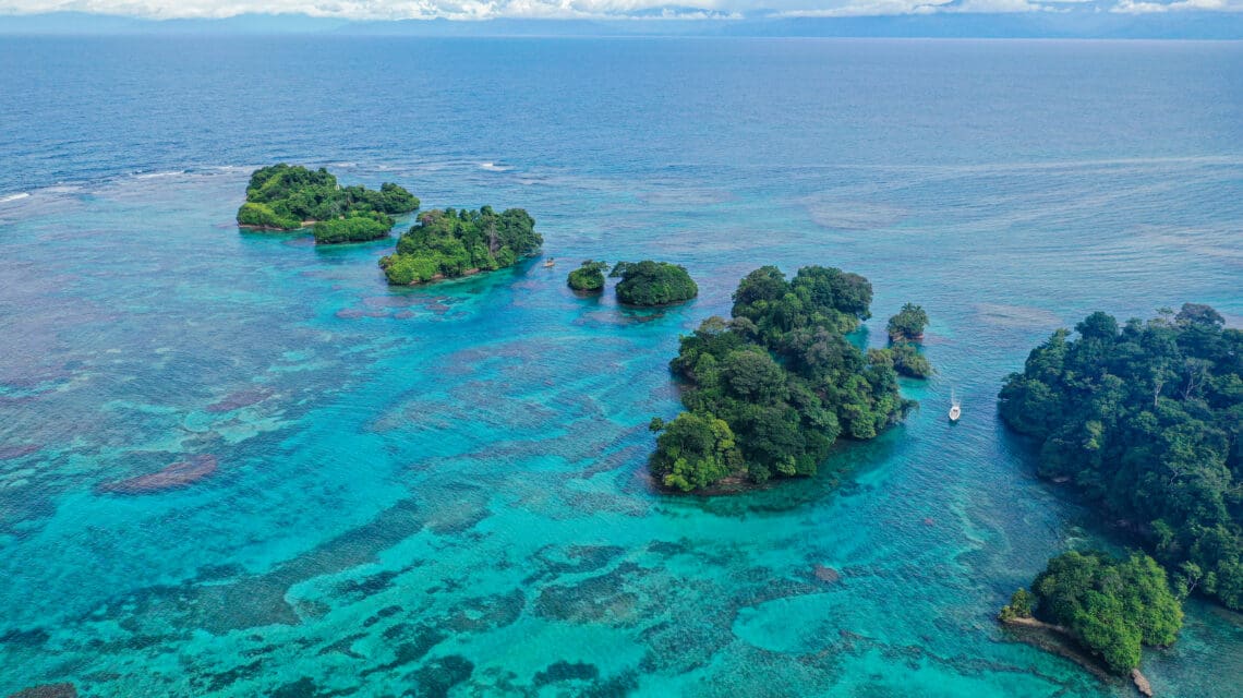 Aerial view of small, lush green islands surrounded by clear turquoise water, a lone boat nearby. The serene scene contrasts with the vast blue ocean.