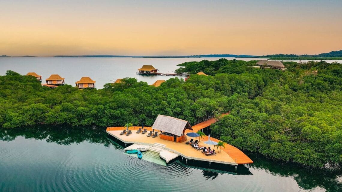 Aerial view of a tropical resort with stilted cabins over water, lush greenery, and a wooden deck with sun loungers. The sky is warm, evoking tranquility.