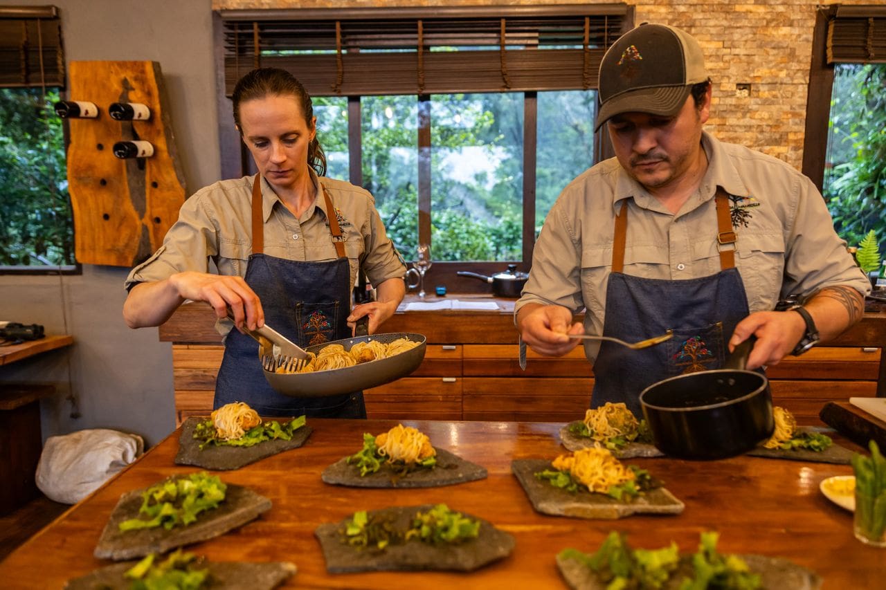 Two chefs in aprons artfully plate dishes in a rustic kitchen, surrounded by greenery outside. The scene conveys focus and culinary artistry.