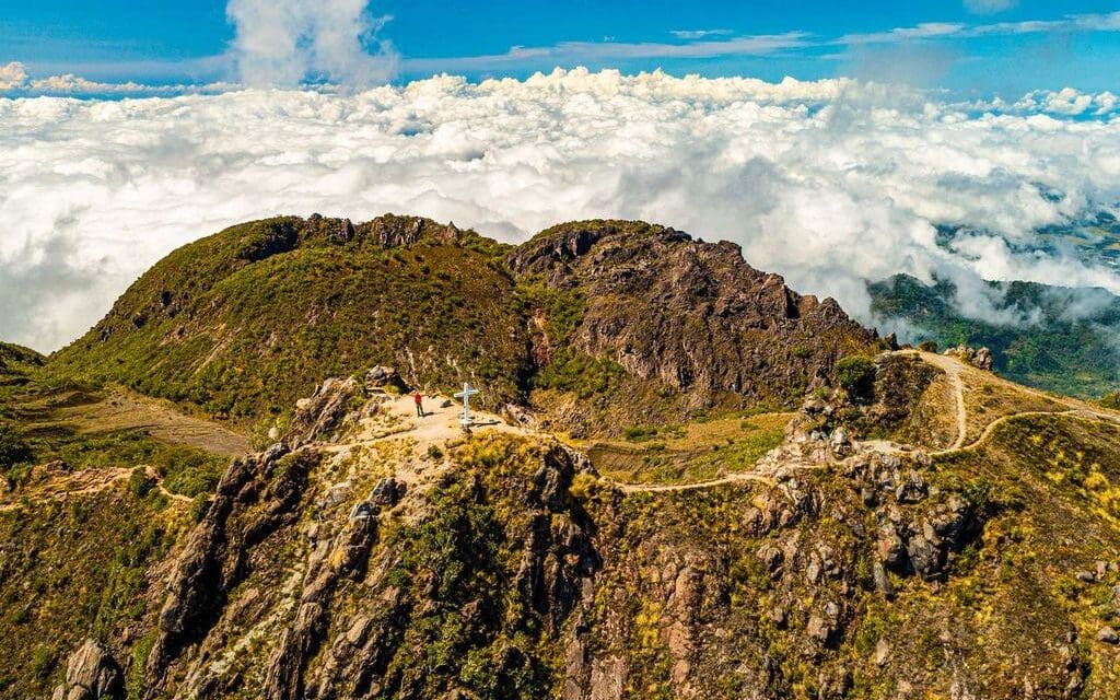 Aerial view of a mountain ridge with a path leading to a cross at the summit. Lush greenery covers the slopes, and fluffy clouds fill the horizon under a clear blue sky.