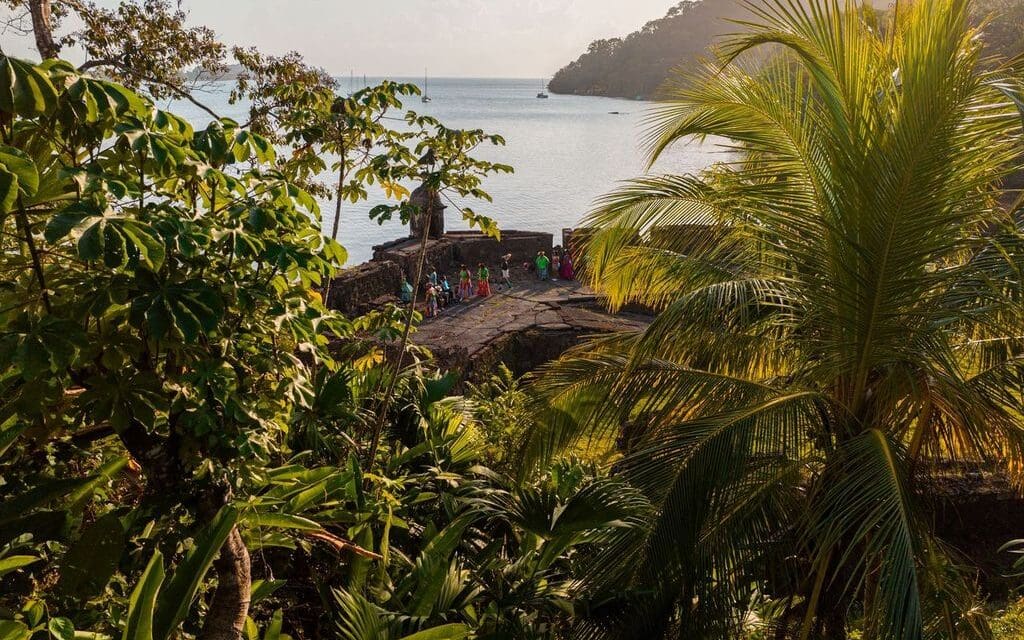 Lush green jungle overlooking a stone fort by the sea, with people in colorful clothing exploring. The sun casts a warm glow over the peaceful scene.