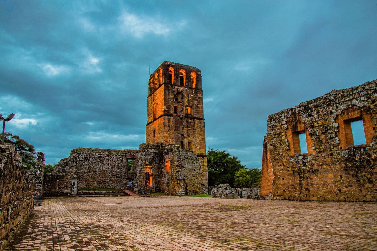 Ancient stone ruins under a dramatic cloudy sky at dusk, with an illuminated tower glowing orange, creating a mysterious and historic ambiance.
