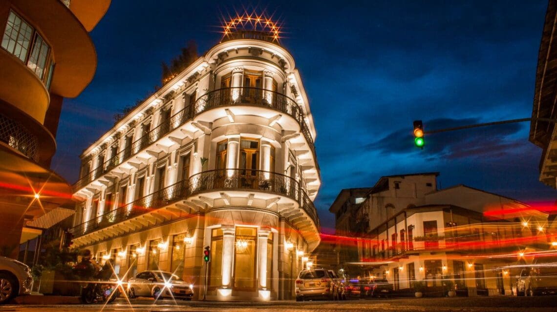 Historic three-story building with ornate balconies, warmly lit at night. Surrounding streaks of car lights add movement under a deep blue sky.