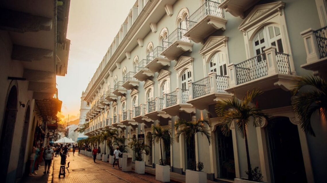A historic street at sunset features an elegant white building with ornate balconies. Palm trees line the sidewalk, and people stroll leisurely.
