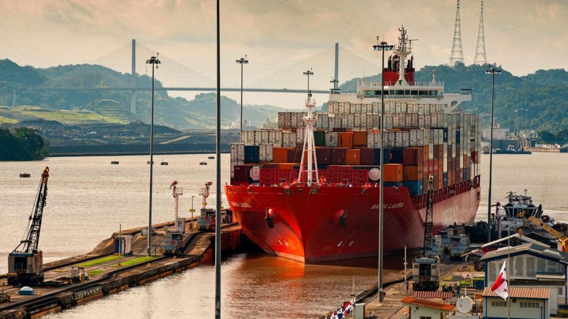A large red cargo ship with colorful containers passes through a canal lock under a cloudy sky, with hills and a bridge in the background.