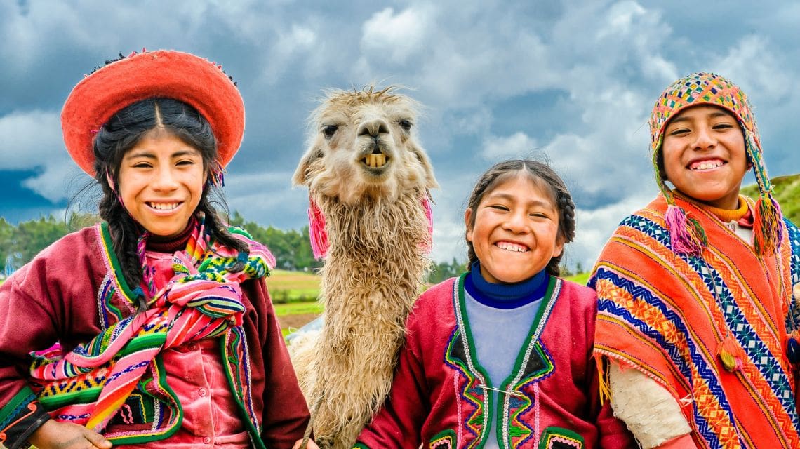 Three smiling children in colorful traditional clothing pose with a llama against a backdrop of a cloudy sky and green landscape, conveying joy and culture.