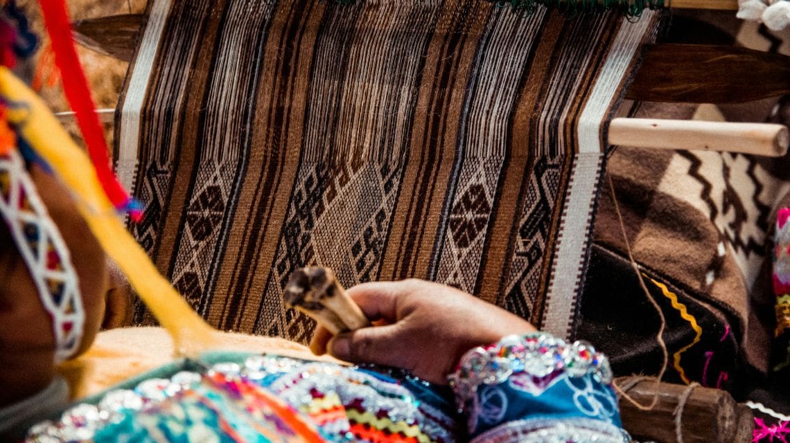 A person in colorful traditional attire weaves on a loom with intricate brown and white patterns. The scene conveys cultural craftsmanship and focus.
