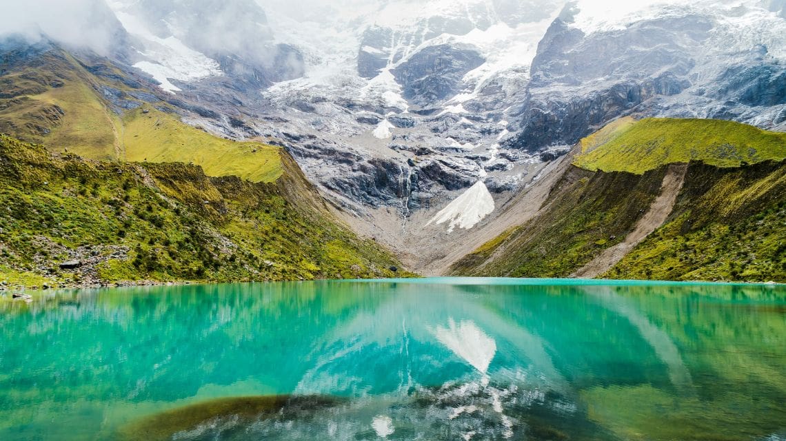 A serene turquoise lake reflects surrounding snow-capped mountains and lush green hills under a cloudy sky, conveying tranquility and natural beauty.