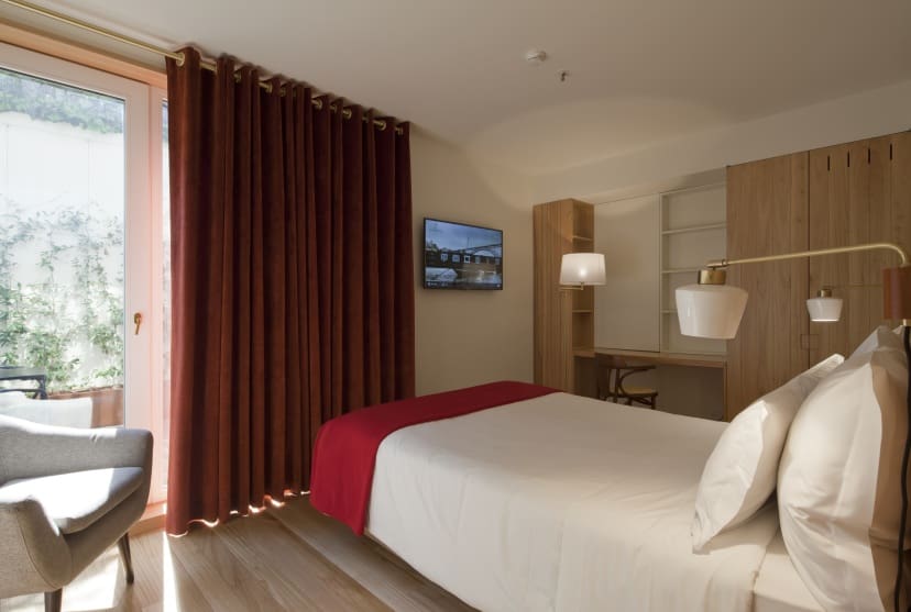 Modern hotel room with a neatly made bed, red blanket, and soft lighting. Curtains cover a window. A desk, chair, and TV create a cozy ambiance.