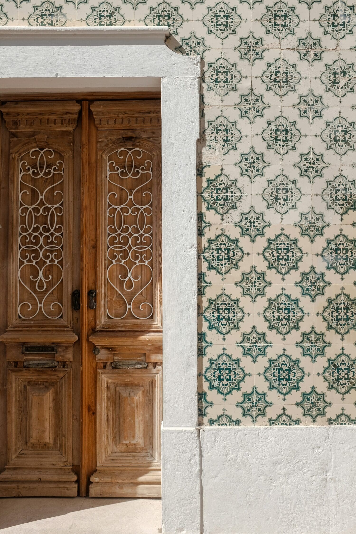 Ornate wooden doors with intricate white ironwork are set against a wall of vintage green and white patterned tiles. The scene exudes a classic, rustic charm.
