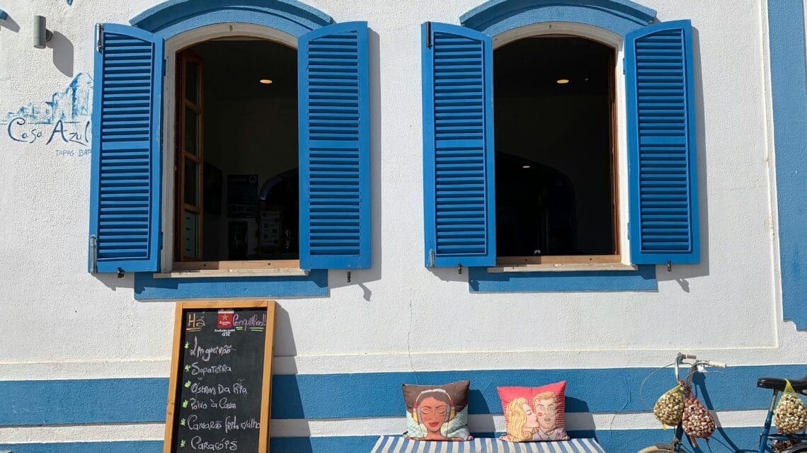 White building with two blue-shuttered windows. Below, a bench with colorful pillows and a menu board. A vintage bike leans nearby, adding a quaint charm.