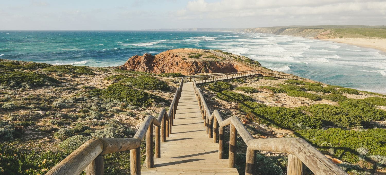 Wooden boardwalk leads to a rocky coastline with waves crashing on the shore, surrounded by green shrubs under a partly cloudy sky. Serene and scenic.