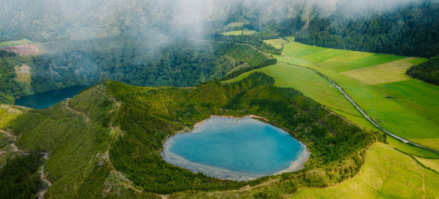 Aerial view of a serene lagoon surrounded by lush greenery and rolling hills. A small blue lake nestles in the foreground, under a partly cloudy sky.