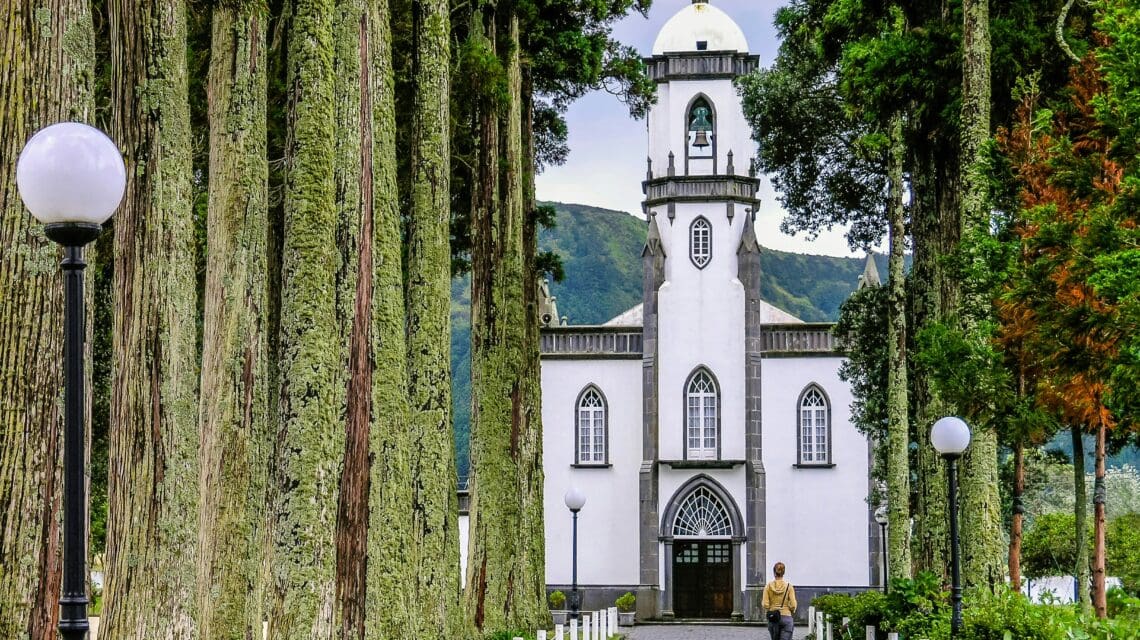 A white church with a central tower stands at the end of a path lined with tall, symmetrical trees and lampposts, exuding a peaceful and serene atmosphere.