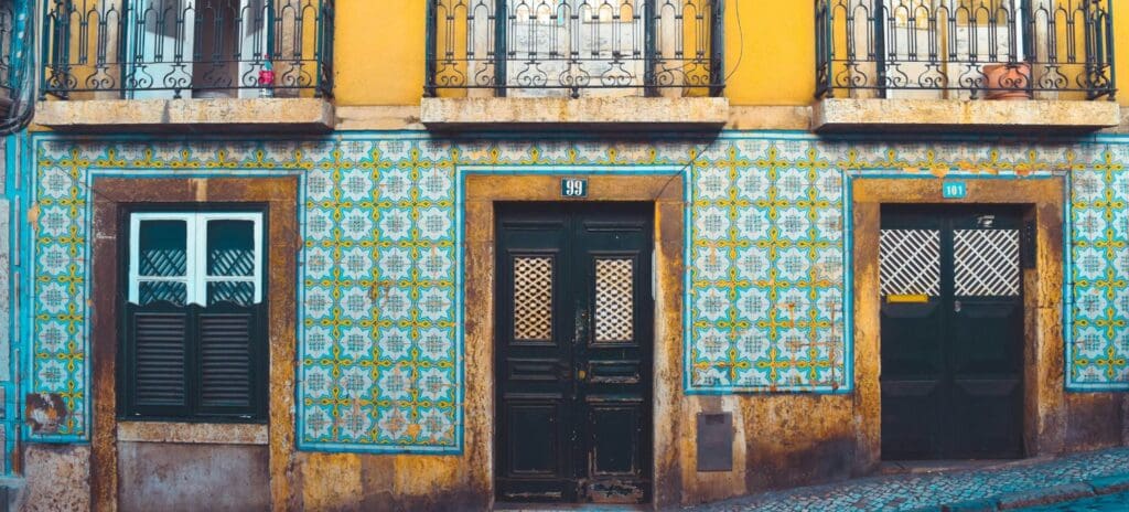 Ornate building facade with intricate blue and yellow tiles, two dark wooden doors, shuttered windows, and wrought iron balconies above, evoking a vintage charm.