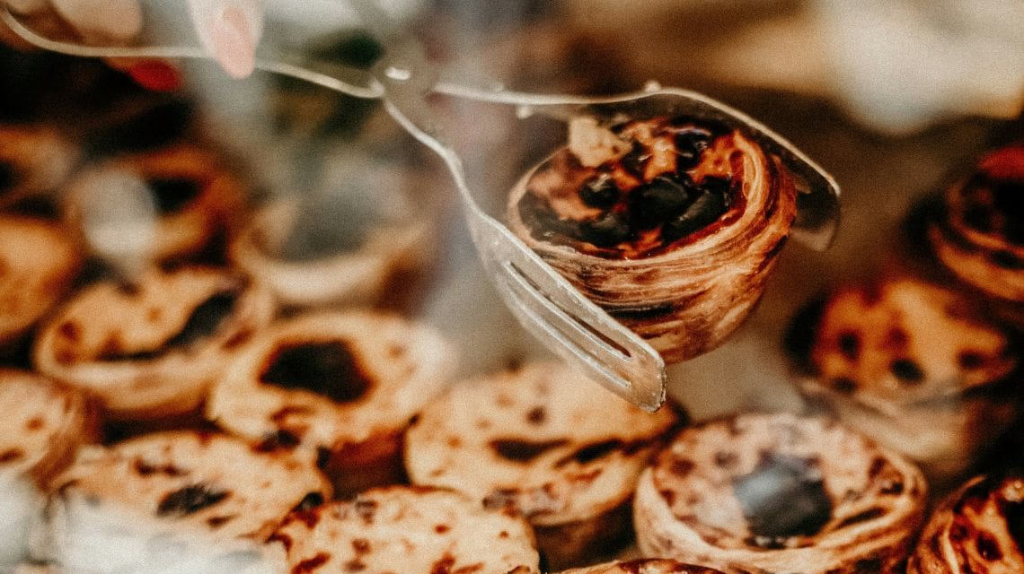 A close-up image of a metal tong holding a single, golden-brown pastry, surrounded by a blurred background of similar pastries, conveying warmth and indulgence.