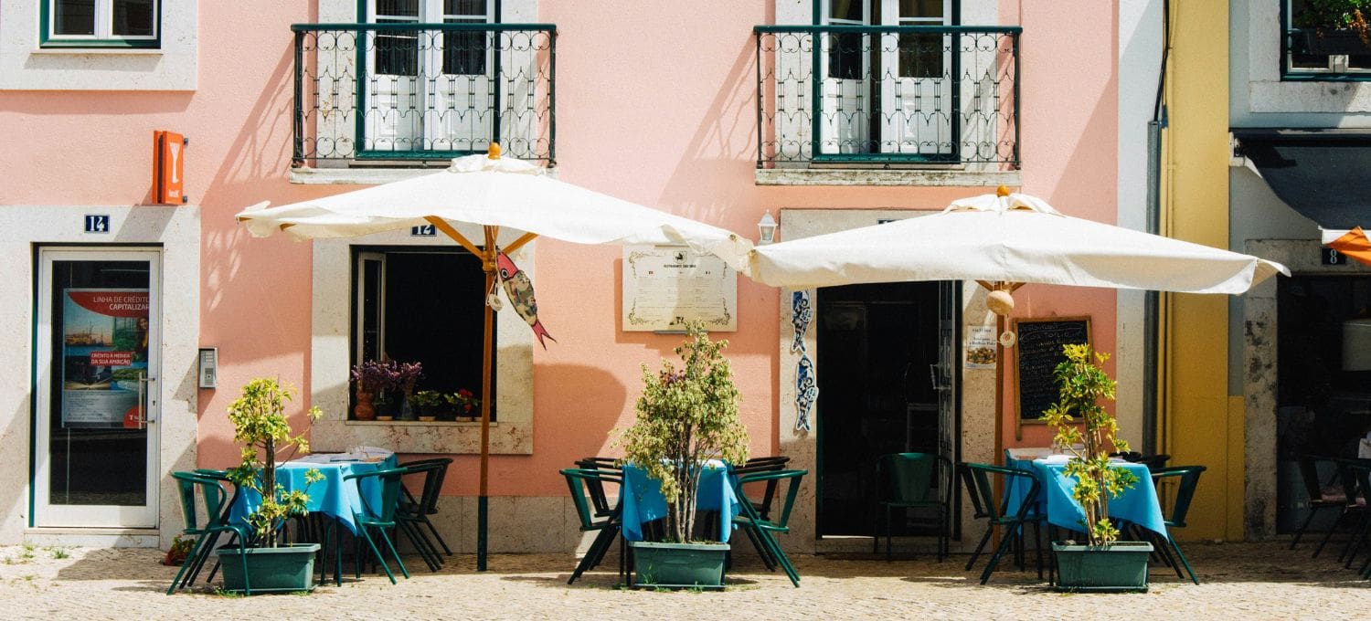 Outdoor café in front of a pink building with two white umbrellas shading tables with blue cloths. Potted plants add a vibrant touch. Relaxed, sunny atmosphere.