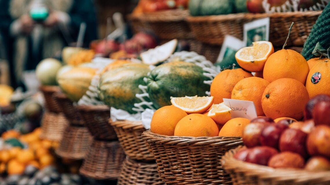 Vibrant farmers market display with baskets of oranges, apples, and tropical fruits. Background shows two blurry figures, evoking a lively, fresh atmosphere.