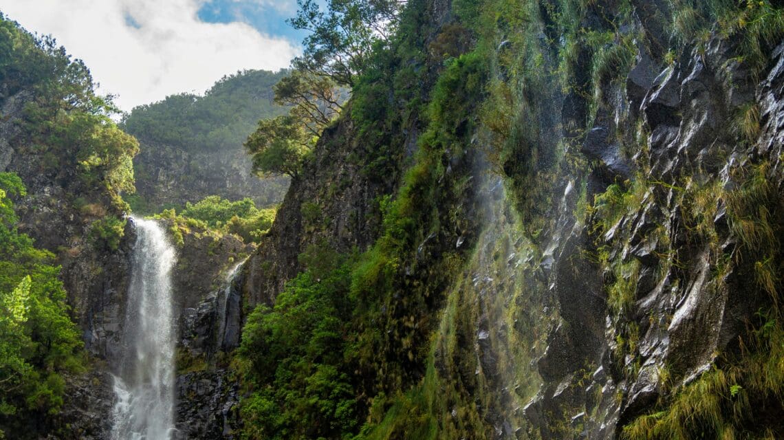 A waterfall cascades down a lush, green cliff under a partly cloudy sky. Mist rises from the falls, adding a serene, natural atmosphere.