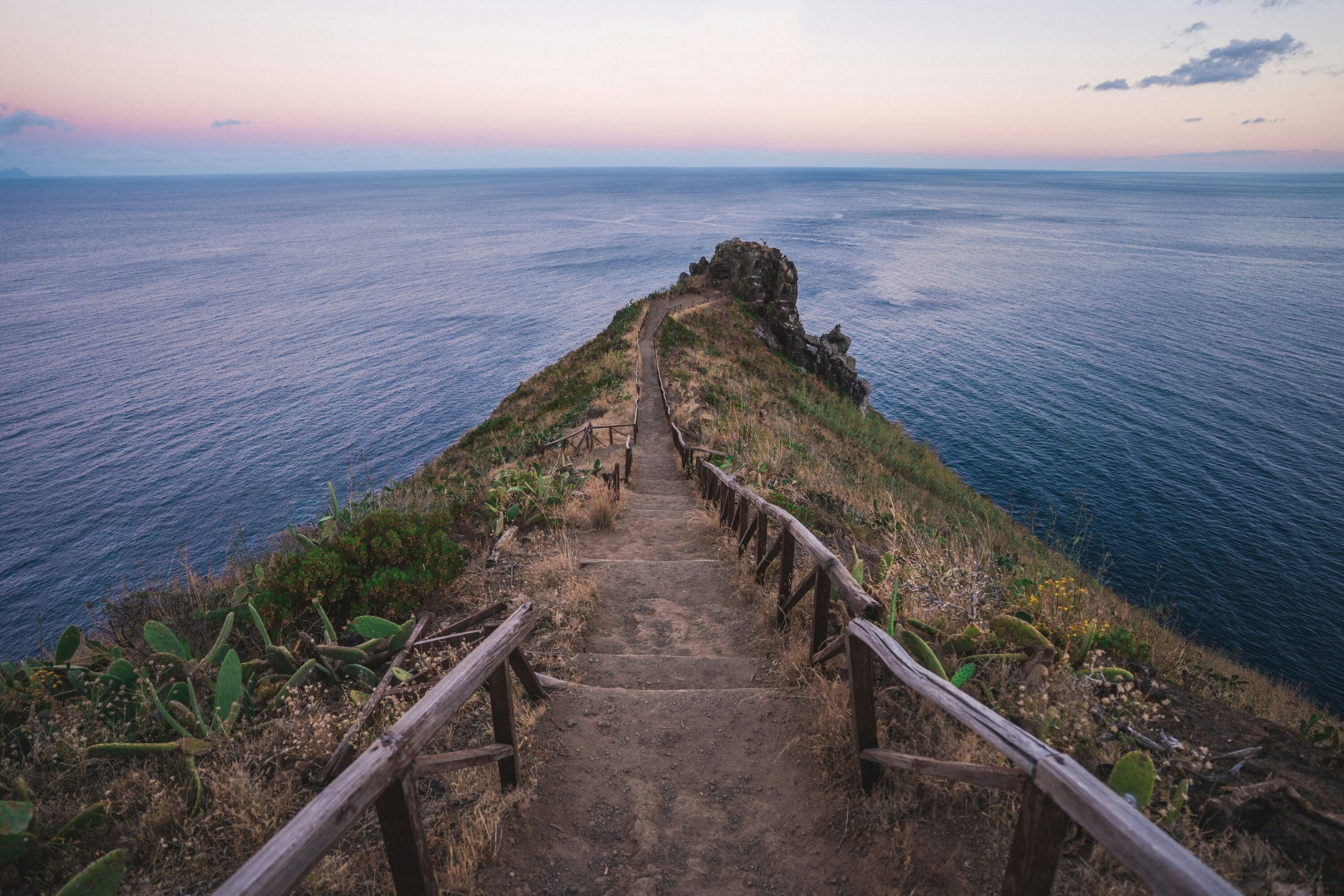 Narrow dirt path flanked by wooden railings leads to a cliff's edge overlooking a vast ocean. The sky is pastel at sunset, evoking tranquility.
