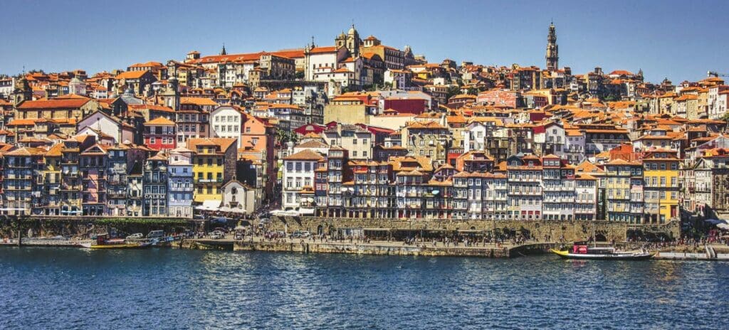 A vibrant coastal cityscape featuring colorful, historic buildings with terracotta roofs lining the waterfront. The scene is lively under a clear blue sky.