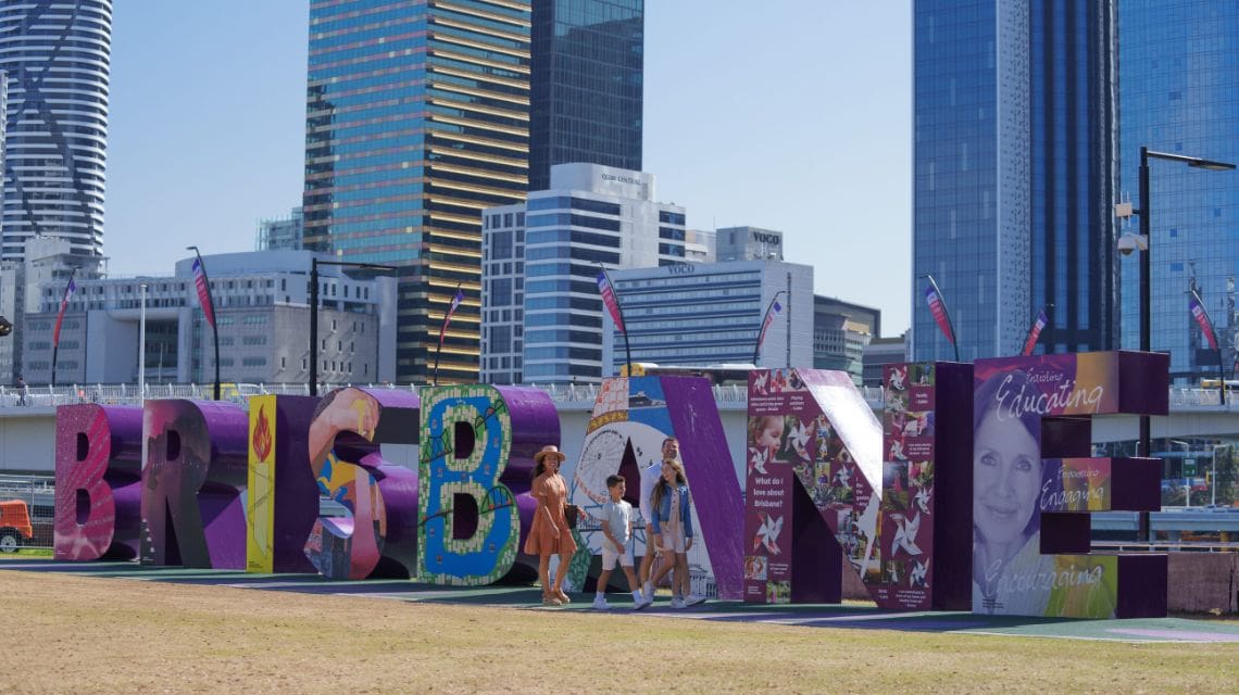 Colorful Brisbane sign with people walking in front, set against a backdrop of modern city skyscrapers under a clear blue sky