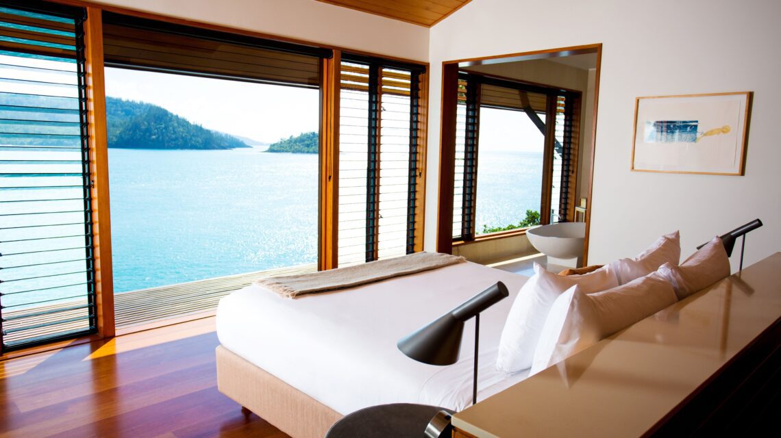 Luxurious bedroom with large windows offering stunning ocean views. The room features a cozy bed, sleek decor, and warm wooden flooring, creating a serene vibe.