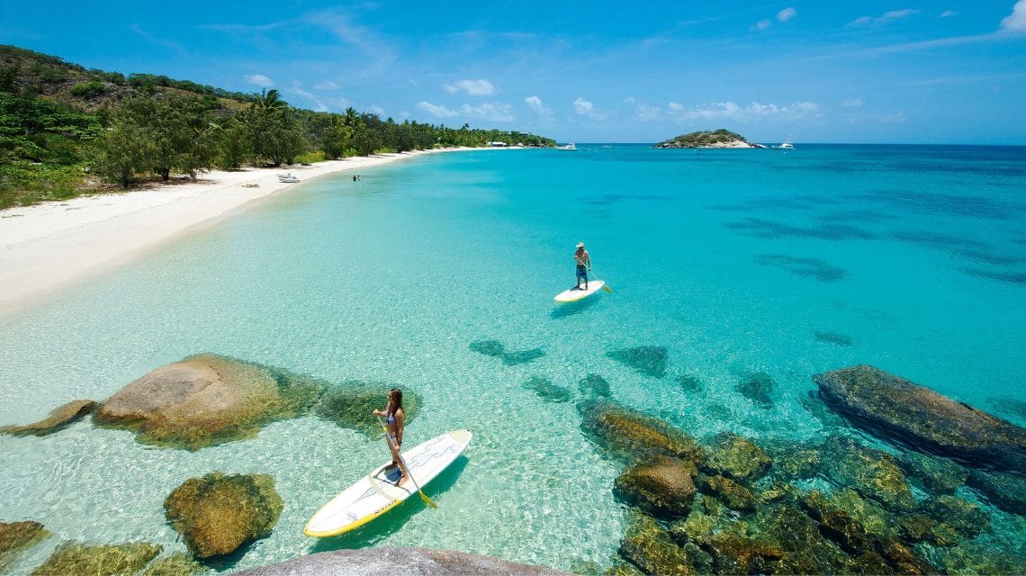 Two people paddleboarding on crystal-clear turquoise water near a sandy beach with lush greenery and rocks. The atmosphere is serene and tropical.